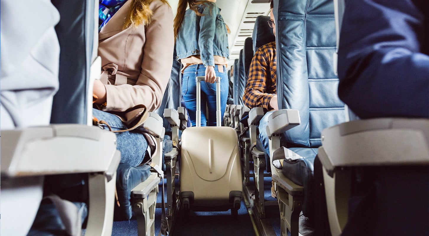 Inside an airplane cabin, passengers are seated while one traveler pulls a suitcase down the aisle searching for a seat.
